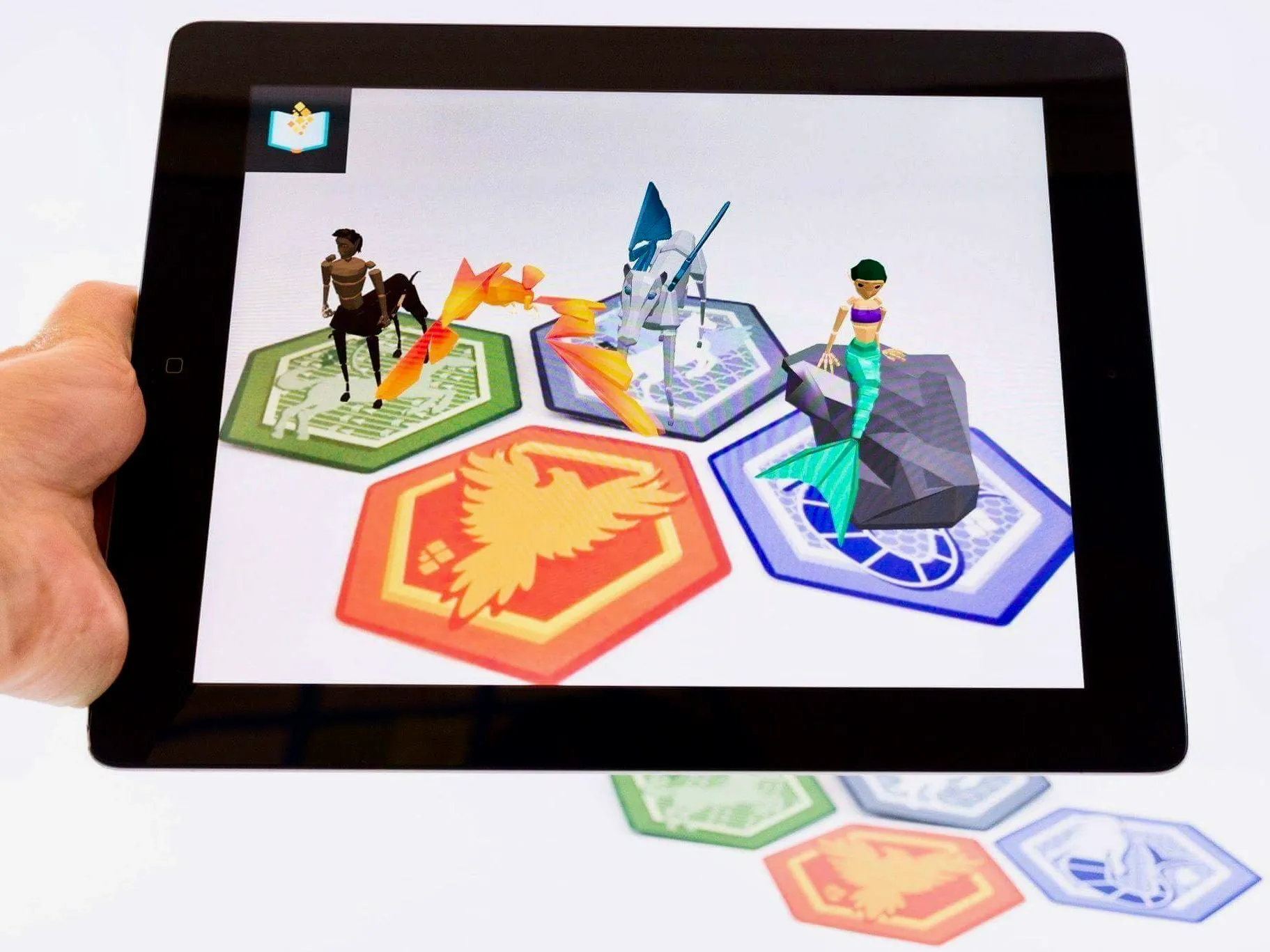 A tablet held over four hexagonal cards, three of them sprouting a creature on screen — a centaur, a horned horse, and a mermaid