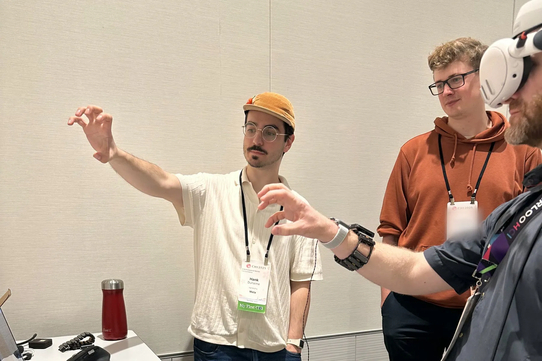 Hank mid-gesture explaining the demo at a conference table, with an attendee in a VR headset and haptic wristband trying it alongside