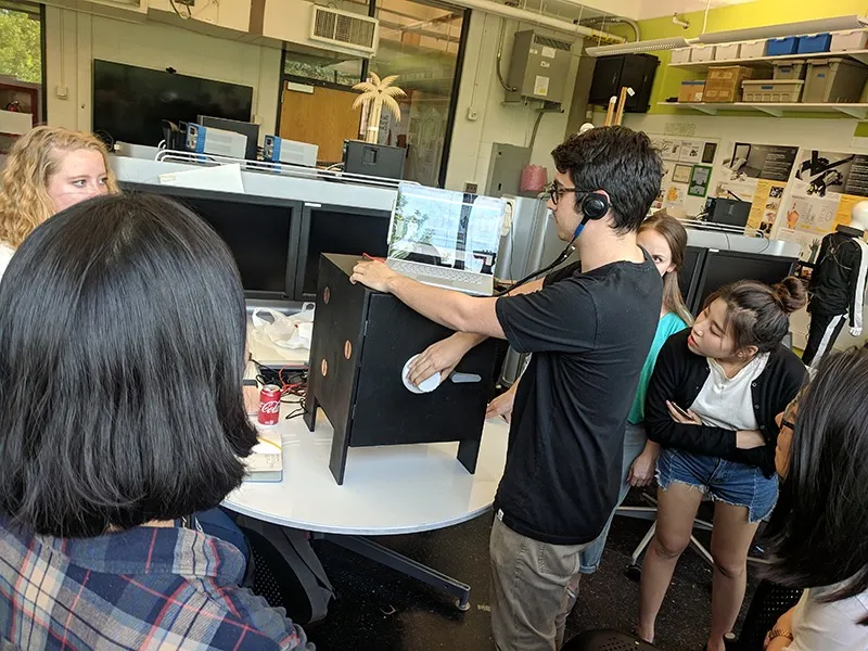 A player listening through the stethoscope at a touch point on the boxy prototype while a group watches