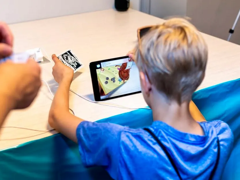 A boy holds a printed card up to a tablet showing the shipwreck scene while a clinician works on his other arm