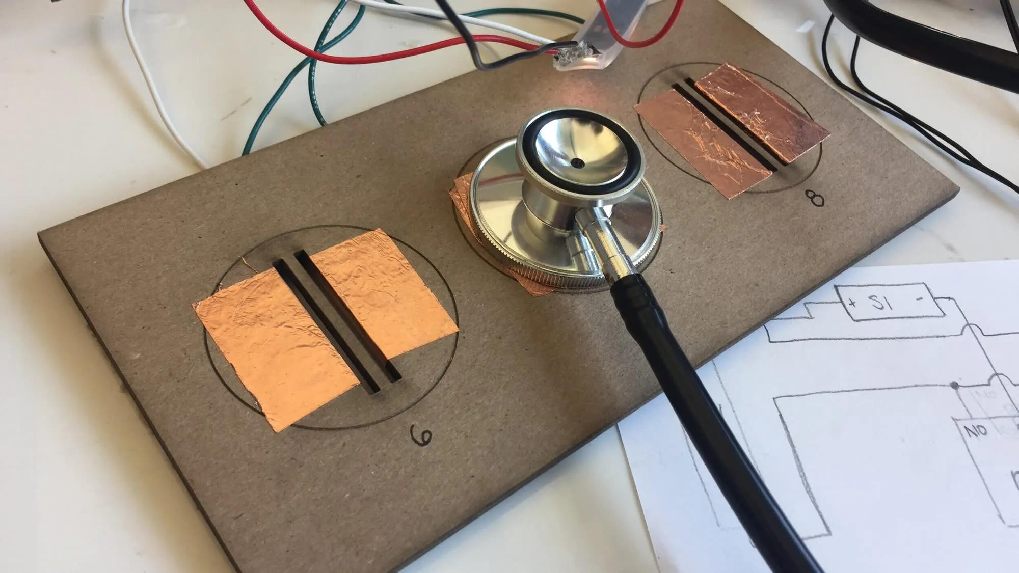 Copper pads taped to a board with a stethoscope resting on one, wired to a breadboard beside a hand-drawn circuit