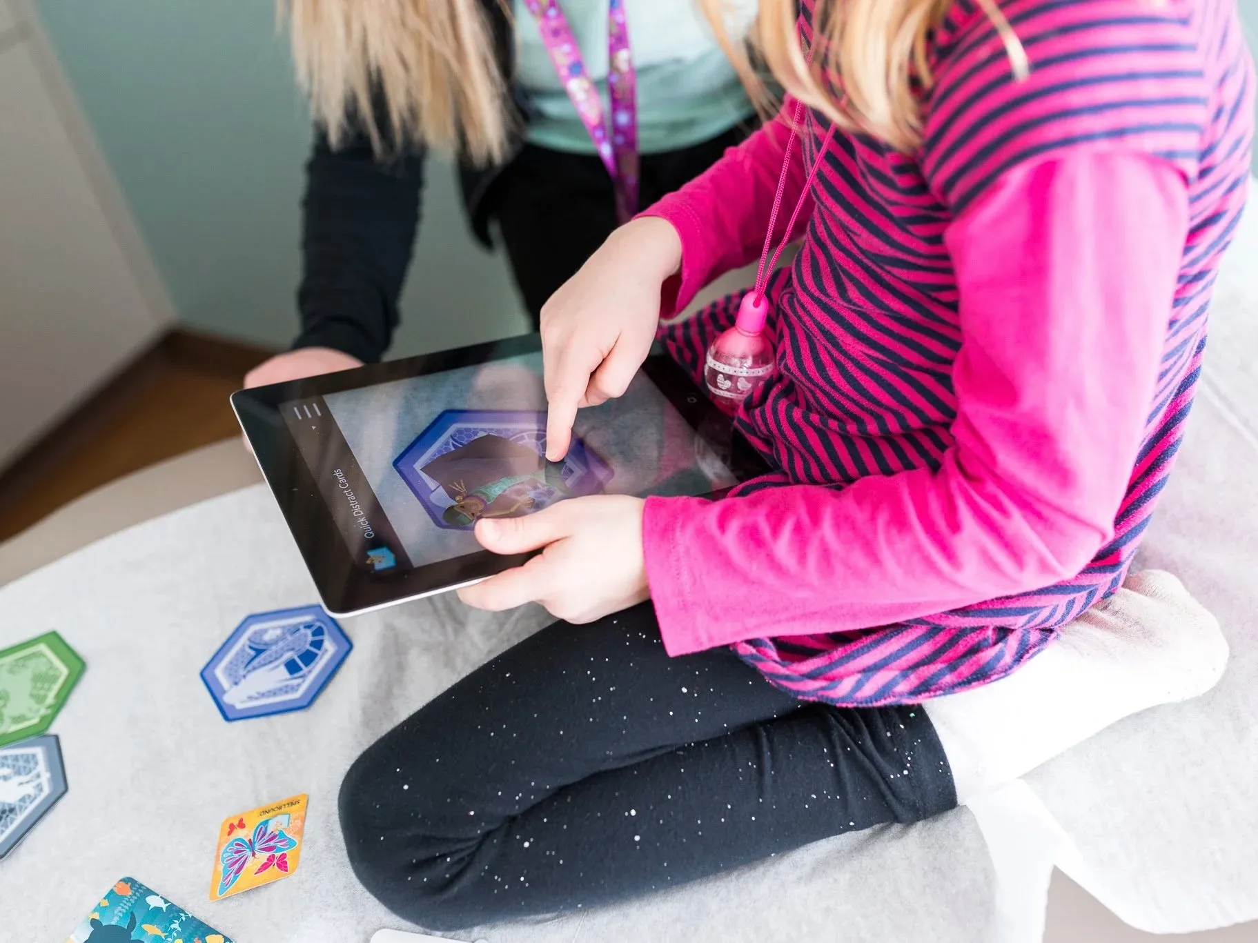 A child sitting cross-legged on a hospital bed taps a tablet showing a mermaid card, more cards scattered beside her